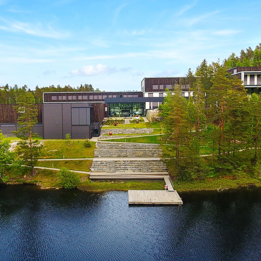 Bilde av skolen fra utsiden. Vann på siden og skog rundt. 